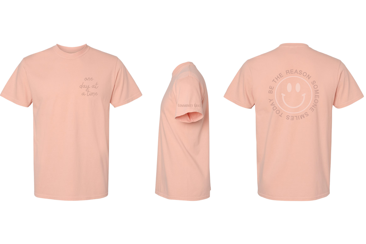 peach short sleeve shirt with "one day at a time" on the left chest area of the shirt and Community Grace on the sleeve in dark orange tones. On the back of the shirt there is a smiley face in a pale peach color with the words "BE THE REASON SOMEONE SMILES TODAY" around it in capital letters in a darker orange tone