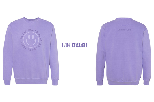 violet crewneck sweatshirt with a smiley face in a vibrant lavender tone with the words "BE THE REASON SOMEONE SMILES TODAY" around it in capital letters in a violet color on the front of the sweatshirt. On the back of the sweatshirt is Community Grace in a violet color. The inner left sleeve of the sweatshirt has the affirmation "I Am Enough"
